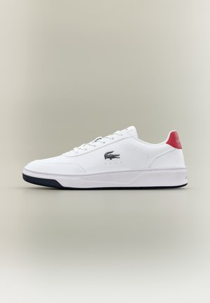 Baskets blanches basses avec semelle noire, logo crocodile noir sur le côté, lacets blancs et languette rouge au talon avec texte de la marque.