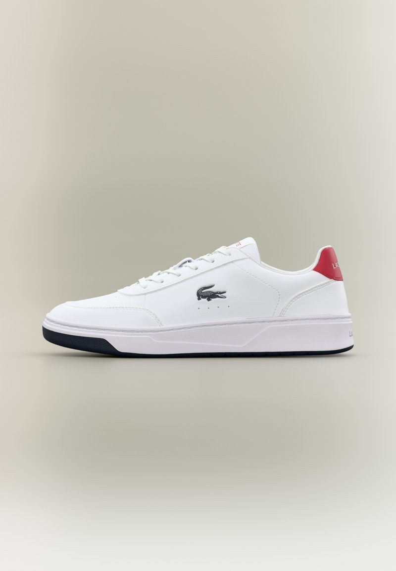Baskets blanches basses avec semelle noire, logo crocodile noir sur le côté, lacets blancs et languette rouge au talon avec texte de la marque.