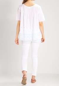 Hvid top med korte ærmer, der har plisseringer bagpå og blå broderet bølget kant. Sættes sammen med hvide slim-fit bukser og beige sandaler.