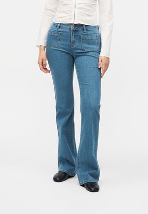 Osoba v modrých bootcut džínsoch s prednými našitými vreckami, bielom tričku s dlhým rukávom a v čiernych topánkach, stojí pred jednofarebným pozadím.