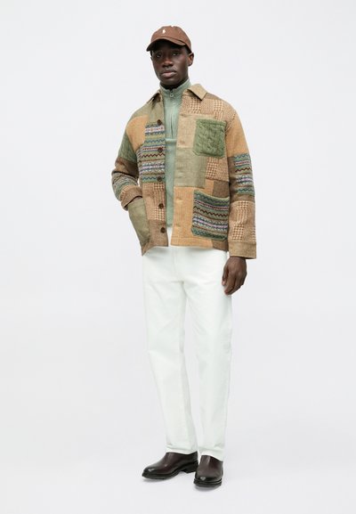 Casaco patchwork em tons terrosos com tecidos texturizados, combinado com gola alta verde, calças brancas e boné castanho, usado com botas pretas.