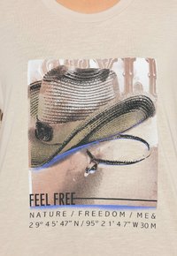 Gráfico de sombrero de vaquero con una pluma y texto "SIENTE LIBRE", "NATURALEZA / LIBERTAD / YO", y coordenadas geográficas sobre tela beige.