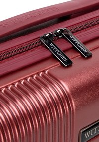 Tuhý kufr v kovově burgundské barvě s ribbovanou texturou. Disponuje dvojitými zipy s černými prvky a s podpisovou rukojetí.