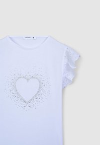 T-shirt bianco in cotone con maniche a volant in pizzo, con un design a forma di cuore realizzato in borchie argentate, centrato sulla parte anteriore. Etichetta della taglia visibile.