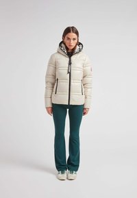 Giacca a vento beige con cappuccio nero con logo, zip frontale e tasche laterali, indossata con pantaloni teal e sneakers color crema.