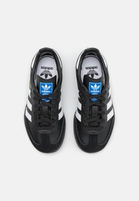 Baskets de sport noires avec trois rayures blanches, lacets noirs, suède noir texturé et un tag bleu Adidas sur la languette.
