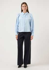 Φωτεινό γαλάζιο πουκάμισο με κουμπιά και φουσκωτά μανίκια με κεντήματα λουλουδιών· μαύρο παντελόνι με φαρδιά πόδια· μαύρες μυτερές γόβες· ουδέτερο φόντο.