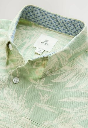 Camisa verde con textura y patrón floral. Forro del cuello en azul claro con un diseño. Dos botones blancos, etiqueta "NEXT" visible. Diseño casual.