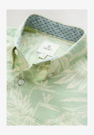 Chemise verte texturée avec un motif floral. Col intérieur à motifs bleu clair. Deux boutons blancs, étiquette "NEXT" visible. Design décontracté.