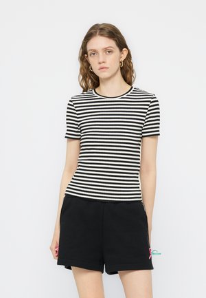 Junge Frau mit welligem braunem Haar, die goldene Creolen-Ohrringe, ein schwarz-weiß gestreiftes kurzärmeliges Oberteil und schwarze Shorts mit kleinen rosa und türkisfarbenen Schleifen trägt.