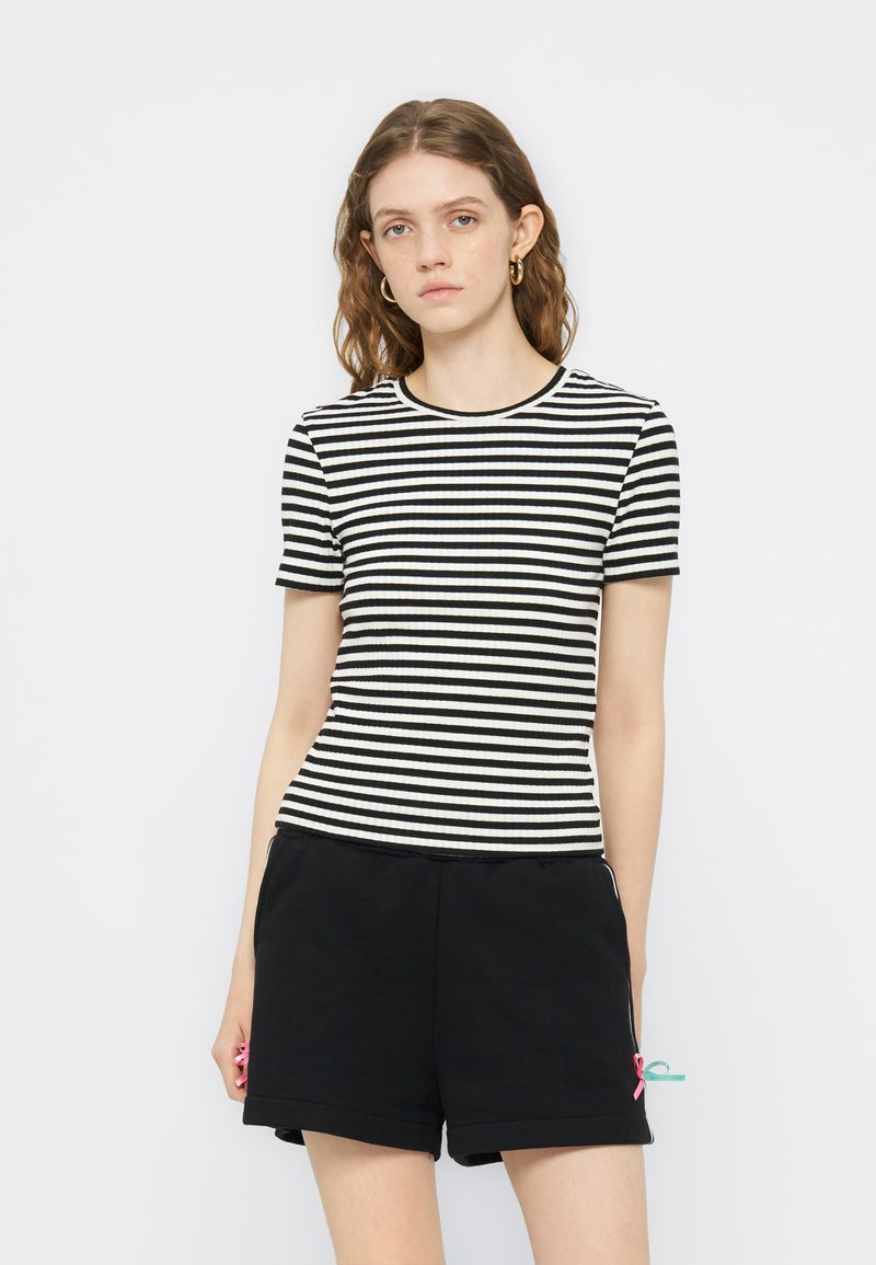 Νέα γυναίκα με κυματιστά καστανά μαλλιά, που φορά χρυσά σκουλαρίκια κρίκους, μαύρο και άσπρο ριγέ κοντομάνικο τοπ και μαύρα σορτς με μικρές ροζ και τιρκουάζ κορδέλες.