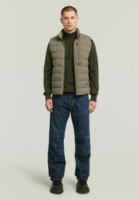 Gilet imbottito oliva con colletto alto indossato sopra un maglione verde scuro, abbinato a jeans blu e scarpe nere, in piedi su uno sfondo pastello.