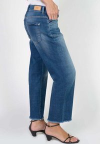 Jean en denim bleu avec une coupe droite, ourlet effiloché, taille mi-haute et détails subtils sur les poches arrière ; portés avec des sandales à talons noires.