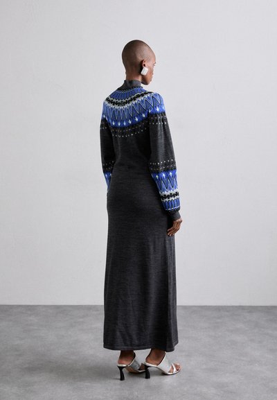 Rochie tricotată cu mâneci lungi, cu un model negru și albastru, având o siluetă ajustată și desene geometrice. Sandale cu toc argintii purtate. Fibră texturată.