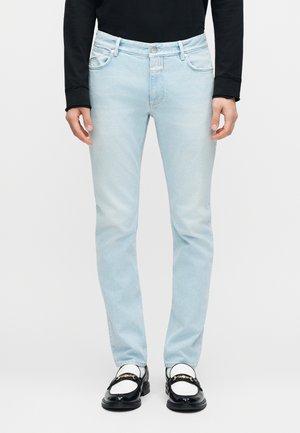 Bărbat purtând blugi slim-fit albastru deschis, pantofi loafer negri cu accente aurii, șosete albe și un pulover negru cu mâneci lungi, pe fundal simplu.