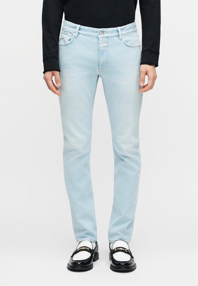 Homme portant un jean slim bleu clair, des mocassins noirs avec des accents dorés, des chaussettes blanches et un haut noir à manches longues sur un fond uni.