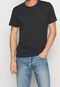 Muž oblečený v černém tričku Levi’s s jemným logem a světle modrými džínami, stojící s jednou rukou v kapse na bílém pozadí.