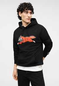 Μαύρο φούτερ με κουκούλα από βαμβάκι, με μεγάλο κόκκινο σκίτσο σκύλου με λευκές λεπτομέρειες. Διαθέτει μπροστινή τσέπη και κουκούλα με κορδόνι.