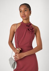 Bordo halter neck obleka z rožnatim aplikejem. Model drži srebrno bleščečo torbico in nosi zlate prstane. Gladek material, prilegajoča silhueta.