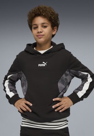 Junge Person mit lockigem Haar, die einen schwarzen Puma-Hoodie mit gemusterten Ärmeln trägt, die Hände in die Hüften gestützt, stehend vor einem grauen Hintergrund.