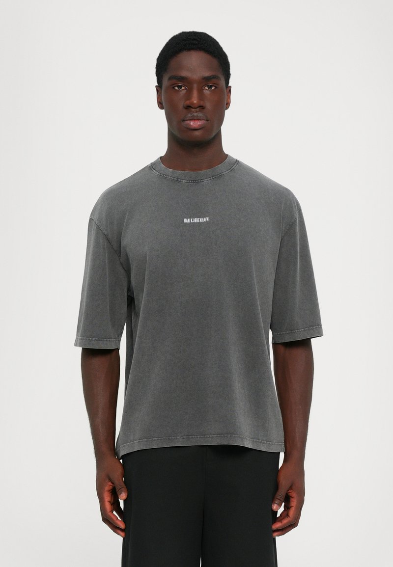Man in een vervaagde donkergrijze oversized t-shirt met korte mouwen en kleine witte "HAN KJØBENHAVN" tekst op de borst, staand tegen een witte achtergrond.