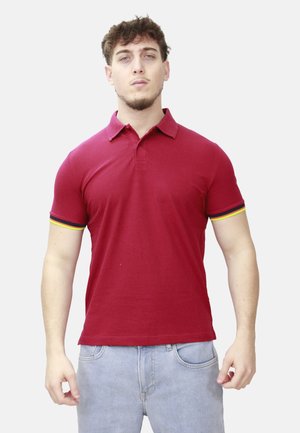Giovane uomo con una polo rossa con strisce gialle e nere sulle maniche e jeans azzurri, in piedi davanti a uno sfondo semplice.