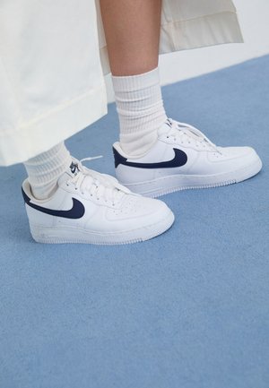 Balti Nike Air Force 1 sneakeriai su tamsiai mėlynais akcentais, perforuota nosimi, lygiais raišteliais ir guminiu padu, derinami su raukšlėtais baltais kojinėmis.