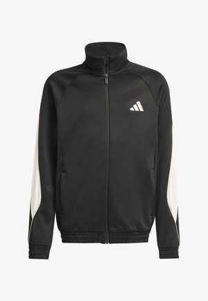 Adolescente ragazzo e ragazza che indossano tute Adidas nere, il ragazzo con cuffie intorno al collo e le braccia incrociate, la ragazza sorridente con una mano in tasca.