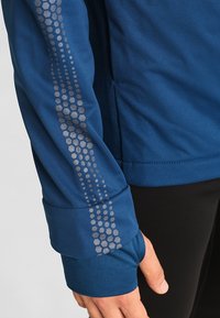 Manica di una giacca sportiva blu con un motivo esagonale riflettente grigio e foro per il pollice, indossata da una persona in pantaloni neri.