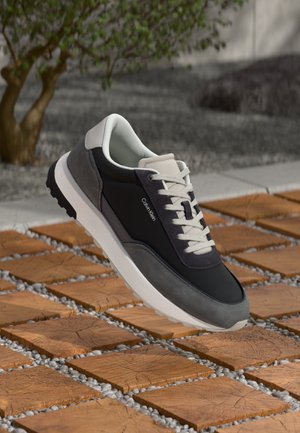 Low-top grey and black Calvin Klein sneaker with white laces floating above wooden block and pebble pathway outdoors.