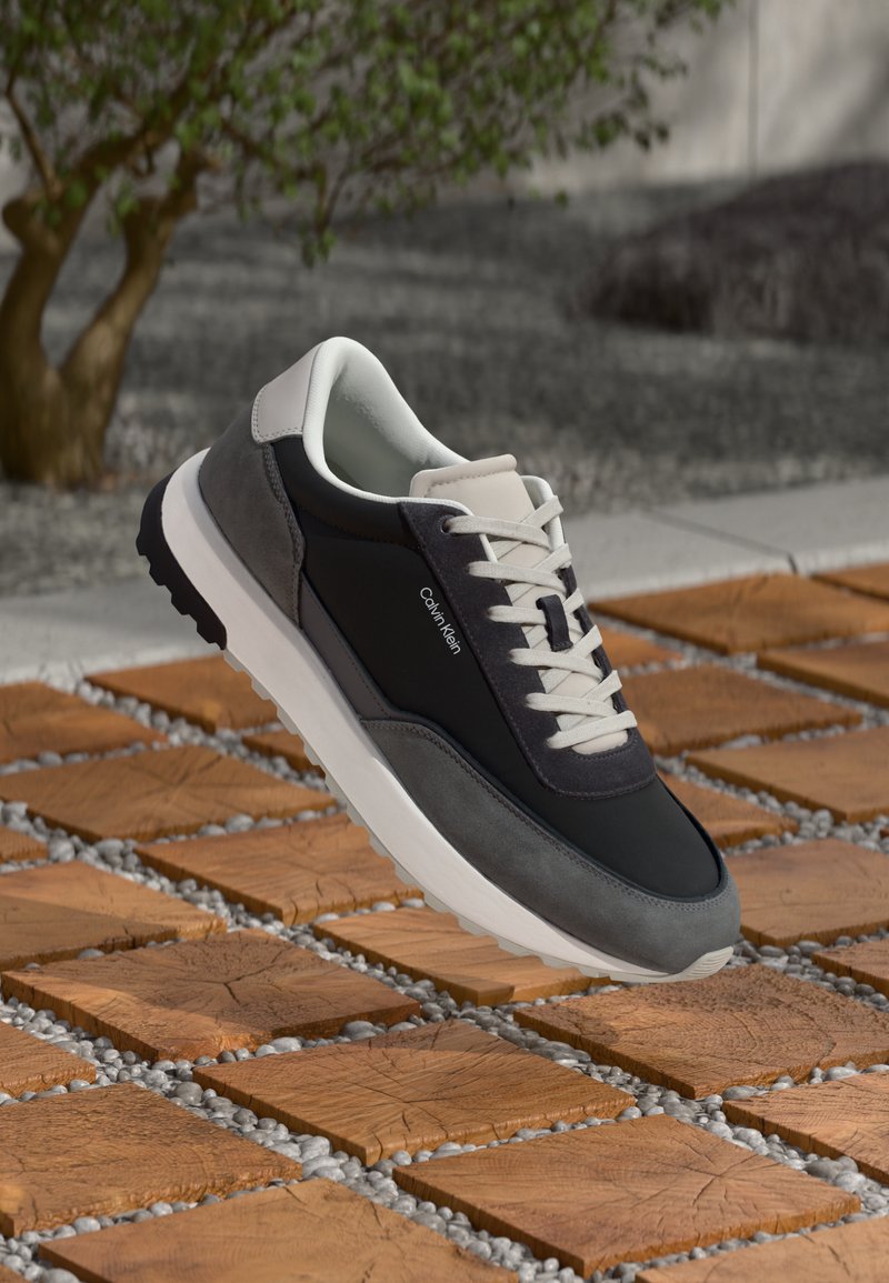 Low-top grey and black Calvin Klein sneaker with white laces floating above wooden block and pebble pathway outdoors.
