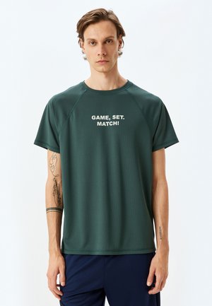 Camisa deportiva de color verde oscuro con mangas cortas y cuello redondo, que presenta el texto blanco "¡JUEGO, SET. PARTIDO!" en la parte frontal. Tejido suave y ligero.