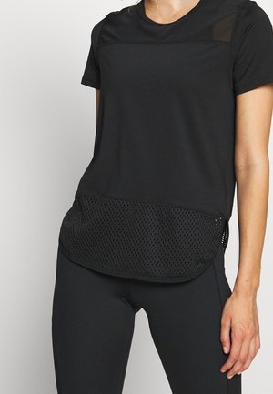 Frau trägt ein schwarzes kurzärmeliges Oberteil mit Mesh-Detail am unteren Vorderteil und schwarze eng anliegende Hose, steht vor einem schlichten Hintergrund.
