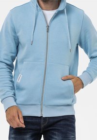 Svetle modrá mikina na zip s přední kapsou, kapucí se šňůrkou, žebrovanými manžetami a malou logo nášivkou na boku. Měkká textilie.