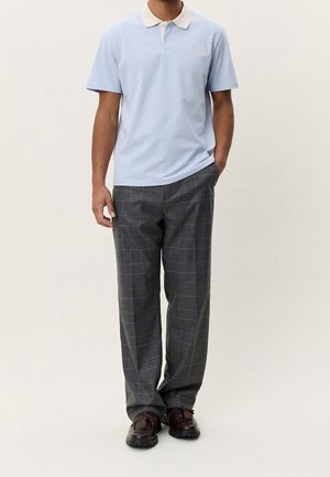 Homme portant un polo bleu clair à manches courtes, un pantalon à carreaux gris et des chaussures en cuir marron foncé, une main dans la poche.