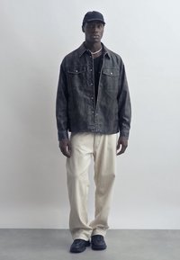 Νεαρός άνδρας που φοράει μαύρο καπέλο μπέιζμπολ, σκούρο τζιν μπουφάν, μαύρο πουκάμισο, κολιέ με μαργαριτάρια, μπεζ παντελόνι και μαύρα αθλητικά παπούτσια, στέκεται μπροστά σε απλό φόντο.