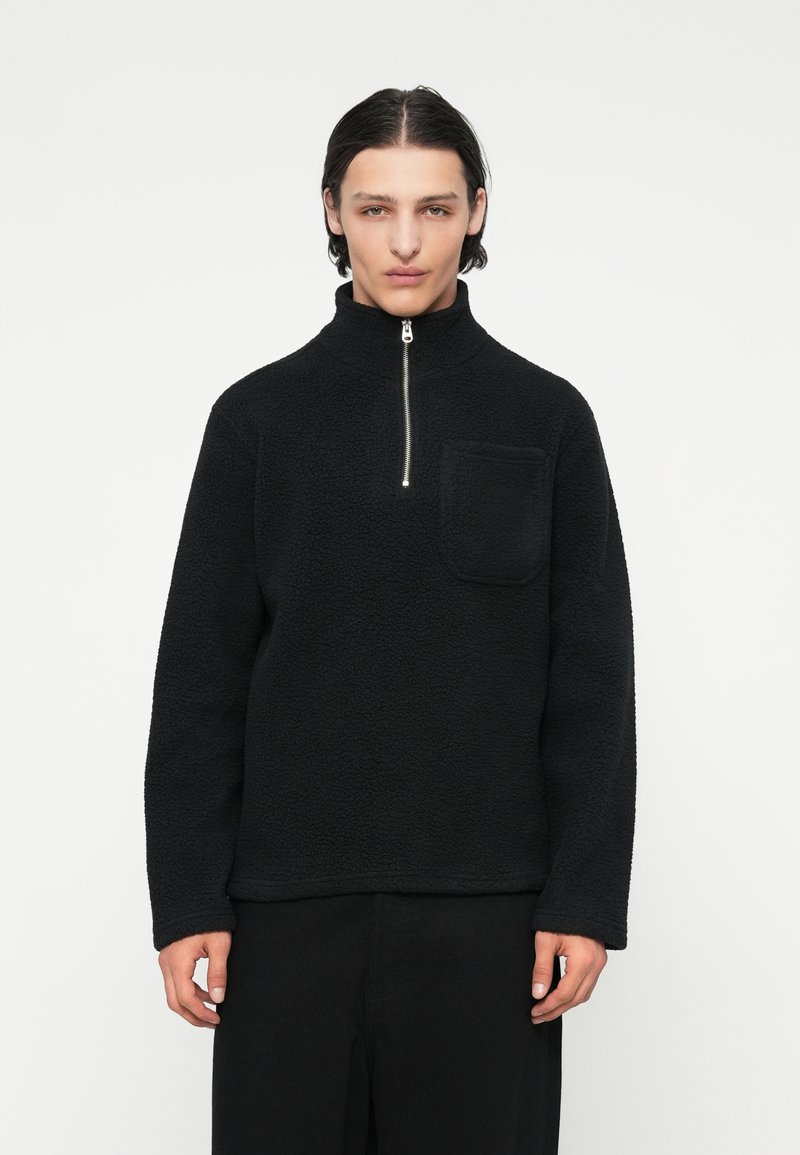 Pulover negru din fleece, cu guler înalt, fermoar până la jumătate și buzunar frontal. Caracteristici includeț țesătură texturată și croială lejeră. Mâneci lungi și design minimal.