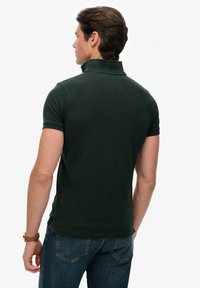 Donkergroen poloshirt met een geribbelde textuur en korte mouwen, voorzien van een omgeslagen kraag en een rechte zoom. Draagt bij een spijkerbroek.