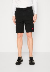 Man i svarta cargoshorts med dragkedjefickor, svarta läder snörskor och en vit långärmad skjorta mot en enfärgad bakgrund.