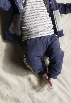 Survêtement bleu marine avec une veste zippée et des accents beige, associé à un T-shirt gris rayé et un pantalon foncé avec un détail en bloc de couleur.