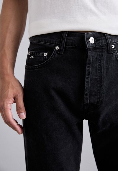 Black denim jeans with silver hardware, flat pockets, and a subtle logo detail; smooth texture and fitted design visible around waist and thighs.