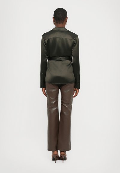 Camicia nera in satin a maniche lunghe cintata in vita, abbinata a pantaloni in pelle marrone scuro a vita alta. Texture liscia, vestibilità su misura.