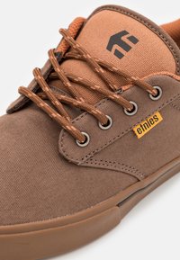 Baskets en toile marron avec des accents orange contrastants, œillets de lacets en métal et logo sur la languette. Dotées d'un tissu texturé et d'une semelle en caoutchouc.