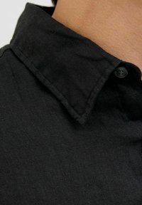 Camisa de lino negra con un cuello estructurado, que presenta un cierre de botones. La tela tiene una textura y un tejido sutil a lo largo.