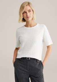 Biały, teksturowany top z krótkim rękawem, z diamentowym wzorem i okrągłym dekoltem, w połączeniu z ciemnymi spodniami w prążki i paskiem.