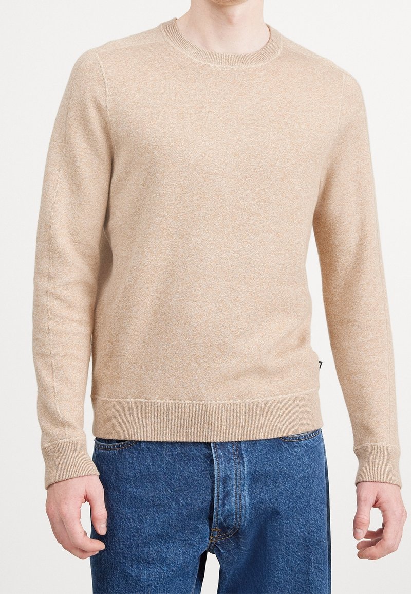 Homme portant un pull beige à manches longues avec col rond et un jean en denim bleu, debout devant un fond clair uni.