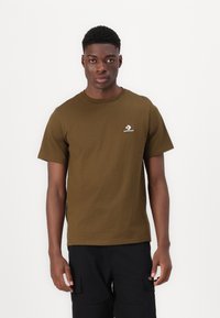 Camiseta de algodón verde oliva con cuello redondo y mangas cortas. Presenta un logo blanco de Converse en el área del pecho izquierdo. Textura suave.