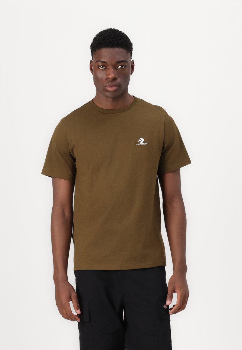 Camiseta de algodón verde oliva con cuello redondo y mangas cortas. Presenta un logo blanco de Converse en el área del pecho izquierdo. Textura suave.