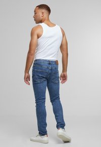 Férfi fehér ujjatlan pólóban, kék farmerben és fehér sportcipőben áll, és balra néz egy sima szürke háttér előtt.