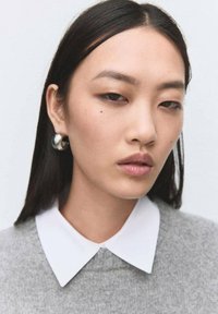 Modelo con cabello negro liso, vistiendo un suéter gris y una camisa blanca con cuello. Luce un pendiente de aro plateado y maquillaje natural.